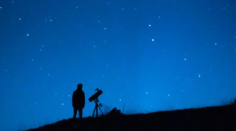 Astronomer observing the immensity of the universe and the stars. Silhouette of a astronomy lover person with a telescope observing the blue starry sky at night.